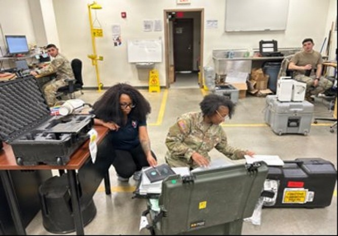 The image shows military personnel and a civilian working in what appears to be a military operations or equipment room. Several individuals are engaged with military-grade equipment cases, documents, and computers. The setting appears to be an indoor facility with standard military workspace characteristics.