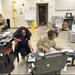 The image shows military personnel and a civilian working in what appears to be a military operations or equipment room. Several individuals are engaged with military-grade equipment cases, documents, and computers. The setting appears to be an indoor facility with standard military workspace characteristics.