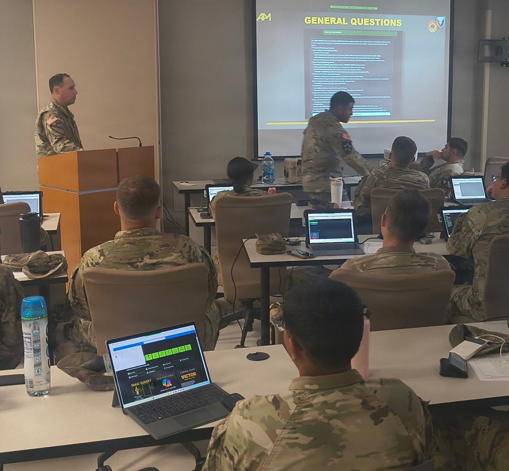 A military classroom training session is underway with multiple soldiers in camouflage uniforms seated at desks with laptops. An instructor in uniform stands at a podium presenting a slide titled 'GENERAL QUESTIONS' on a large projection screen. The setting appears to be a formal military education or briefing environment.