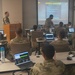 A military classroom training session is underway with multiple soldiers in camouflage uniforms seated at desks with laptops. An instructor in uniform stands at a podium presenting a slide titled 'GENERAL QUESTIONS' on a large projection screen. The setting appears to be a formal military education or briefing environment.