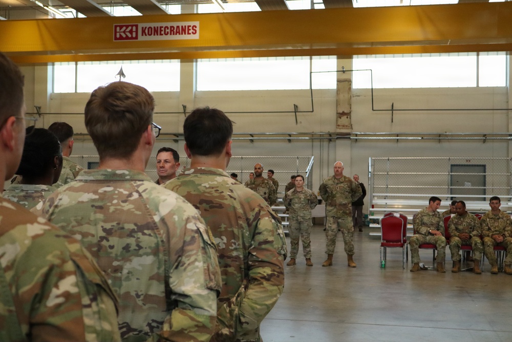 A group of military personnel in OCP (Operational Camouflage Pattern) uniforms are gathered inside a large industrial or maintenance hangar facility. A briefing or address appears to be taking place, with two soldiers standing at the front facing the assembled group. A Konecranes overhead crane system is visible in the background, indicating an industrial or motor pool facility.