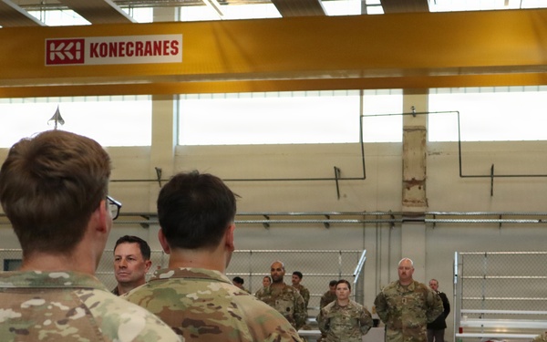 A group of military personnel in OCP (Operational Camouflage Pattern) uniforms are gathered inside a large industrial or maintenance hangar facility. A briefing or address appears to be taking place, with two soldiers standing at the front facing the assembled group. A Konecranes overhead crane system is visible in the background, indicating an industrial or motor pool facility.