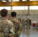 A group of military personnel in OCP (Operational Camouflage Pattern) uniforms are gathered inside a large industrial or maintenance hangar facility. A briefing or address appears to be taking place, with two soldiers standing at the front facing the assembled group. A Konecranes overhead crane system is visible in the background, indicating an industrial or motor pool facility.
