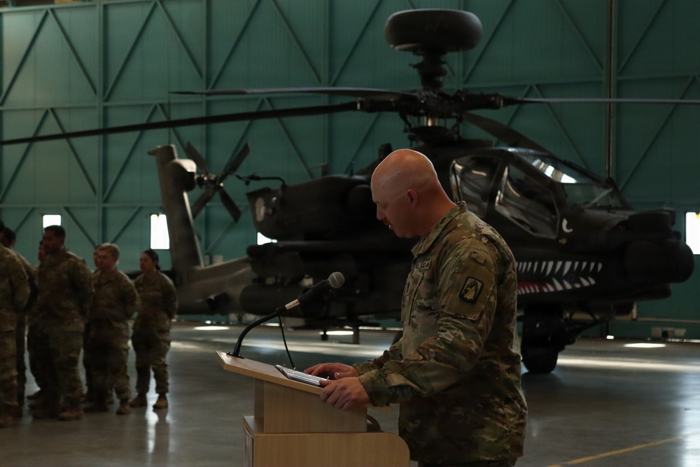 A U.S. Army soldier in MultiCam/OCP camouflage uniform is speaking at a podium inside a military hangar. Behind him is an AH-64 Apache attack helicopter with a distinctive shark mouth nose art. Several other uniformed soldiers are visible standing in the background to the left.