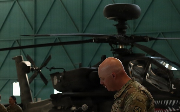 A U.S. Army soldier in MultiCam/OCP camouflage uniform is speaking at a podium inside a military hangar. Behind him is an AH-64 Apache attack helicopter with a distinctive shark mouth nose art. Several other uniformed soldiers are visible standing in the background to the left.