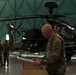 A U.S. Army soldier in MultiCam/OCP camouflage uniform is speaking at a podium inside a military hangar. Behind him is an AH-64 Apache attack helicopter with a distinctive shark mouth nose art. Several other uniformed soldiers are visible standing in the background to the left.