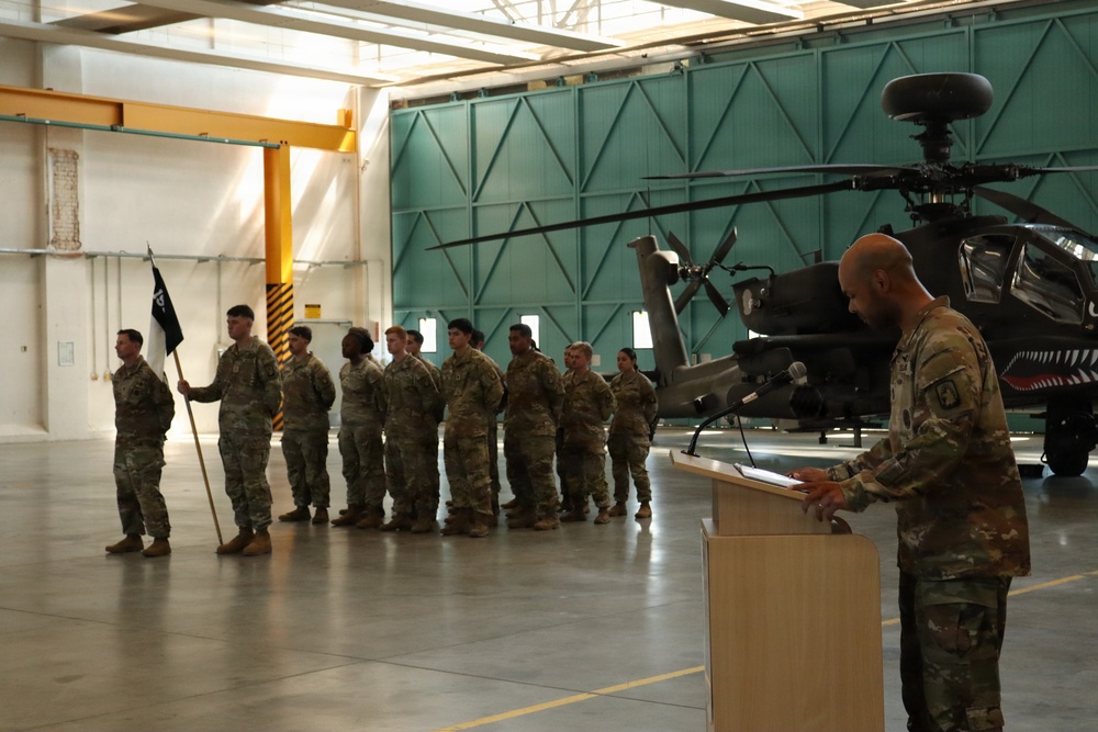 A military ceremony is taking place inside an aircraft hangar, with a soldier speaking at a podium while a formation of soldiers stands at attention holding a unit guidon flag. An Apache attack helicopter is visible in the background, suggesting this is an aviation unit. The setting and uniforms indicate a formal military event such as a change of command, award ceremony, or unit activation.
