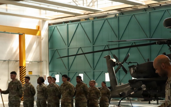 A military ceremony is taking place inside an aircraft hangar, with a soldier speaking at a podium while a formation of soldiers stands at attention holding a unit guidon flag. An Apache attack helicopter is visible in the background, suggesting this is an aviation unit. The setting and uniforms indicate a formal military event such as a change of command, award ceremony, or unit activation.