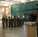 A military ceremony is taking place inside an aircraft hangar, with a soldier speaking at a podium while a formation of soldiers stands at attention holding a unit guidon flag. An Apache attack helicopter is visible in the background, suggesting this is an aviation unit. The setting and uniforms indicate a formal military event such as a change of command, award ceremony, or unit activation.