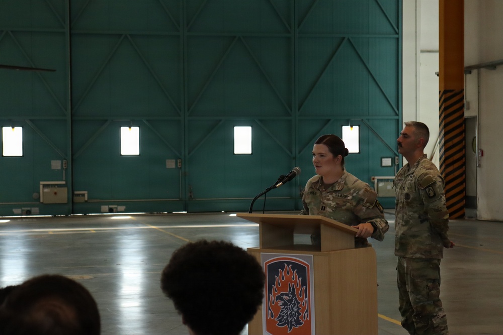 Two U.S. Army soldiers in MultiCam/OCP uniforms are participating in a formal military ceremony inside what appears to be a large aircraft hangar. A female soldier is speaking at a wooden podium bearing a unit insignia patch, while a male soldier stands beside her. The setting suggests an official military event such as a promotion, award, or change of command ceremony.