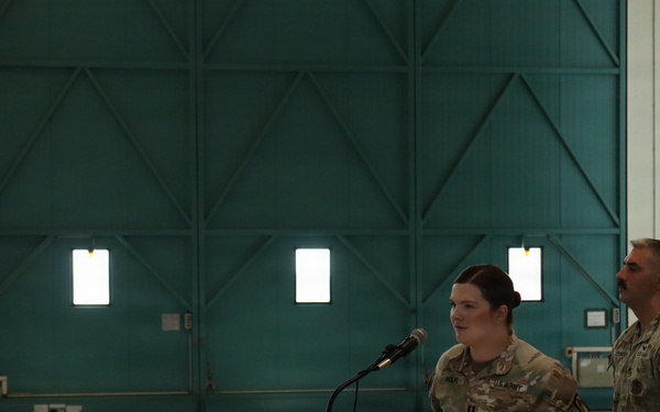 Two U.S. Army soldiers in MultiCam/OCP uniforms are participating in a formal military ceremony inside what appears to be a large aircraft hangar. A female soldier is speaking at a wooden podium bearing a unit insignia patch, while a male soldier stands beside her. The setting suggests an official military event such as a promotion, award, or change of command ceremony.