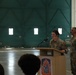 Two U.S. Army soldiers in MultiCam/OCP uniforms are participating in a formal military ceremony inside what appears to be a large aircraft hangar. A female soldier is speaking at a wooden podium bearing a unit insignia patch, while a male soldier stands beside her. The setting suggests an official military event such as a promotion, award, or change of command ceremony.