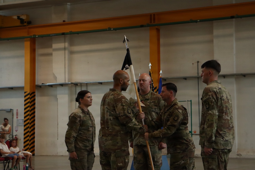 A U.S. Army change of command or activation ceremony is taking place inside what appears to be a military hangar or gymnasium. Several soldiers in OCP (Operational Camouflage Pattern) uniforms are participating in a guidon transfer, with one soldier passing a unit flag (guidon) to another. Civilian family members, including children, are visible seated in the background observing the ceremony.