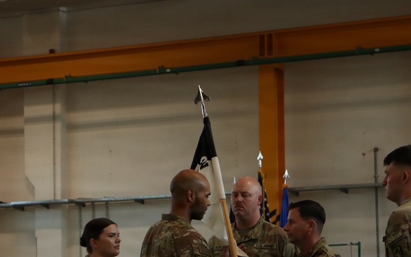 A U.S. Army change of command or activation ceremony is taking place inside what appears to be a military hangar or gymnasium. Several soldiers in OCP (Operational Camouflage Pattern) uniforms are participating in a guidon transfer, with one soldier passing a unit flag (guidon) to another. Civilian family members, including children, are visible seated in the background observing the ceremony.