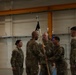 A U.S. Army change of command or activation ceremony is taking place inside what appears to be a military hangar or gymnasium. Several soldiers in OCP (Operational Camouflage Pattern) uniforms are participating in a guidon transfer, with one soldier passing a unit flag (guidon) to another. Civilian family members, including children, are visible seated in the background observing the ceremony.
