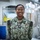 I Keep the Warfighter in the Fight: Hospital Corpsman Second Class Emmanuella Gbogbo A U.S. Navy sailor wearing the Navy Working Uniform (NWU) digital camouflage pattern stands smiling in what appears to be a medical facility or sick bay. The name tape reads 'OGBO' and the rank insignia indicates a Petty Officer Third Class. The background contains medical equipment including an X-ray machine and a shelving unit with medical supplies.