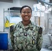 A U.S. Navy sailor wearing the Navy Working Uniform (NWU) digital camouflage pattern stands smiling in what appears to be a medical facility or sick bay. The name tape reads 'OGBO' and the rank insignia indicates a Petty Officer Third Class. The background contains medical equipment including an X-ray machine and a shelving unit with medical supplies.