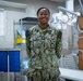 A U.S. Navy service member in NWU Type III (digital camouflage) uniform is standing and smiling in what appears to be a military medical facility or radiology room. The name tape reads 'OGBO' and the branch tape reads 'U.S. NAVY,' with a Petty Officer rank insignia visible on the chest. The background contains medical imaging equipment, including a Siemens X-ray machine, suggesting this is a naval medical treatment facility.