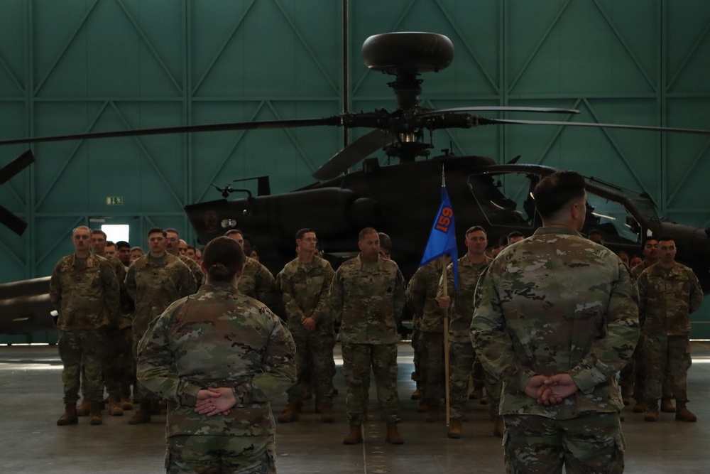 A military formation ceremony is taking place inside a large aircraft hangar. Soldiers in OCP (Operational Camouflage Pattern) uniforms are assembled in formation facing two individuals with their backs to the camera, one of whom holds a blue unit guidon bearing the number '159'. An AH-64 Apache attack helicopter is prominently displayed in the background.