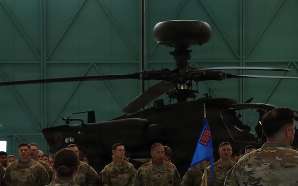 A military formation ceremony is taking place inside a large aircraft hangar. Soldiers in OCP (Operational Camouflage Pattern) uniforms are assembled in formation facing two individuals with their backs to the camera, one of whom holds a blue unit guidon bearing the number '159'. An AH-64 Apache attack helicopter is prominently displayed in the background.