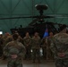 A military formation ceremony is taking place inside a large aircraft hangar. Soldiers in OCP (Operational Camouflage Pattern) uniforms are assembled in formation facing two individuals with their backs to the camera, one of whom holds a blue unit guidon bearing the number '159'. An AH-64 Apache attack helicopter is prominently displayed in the background.