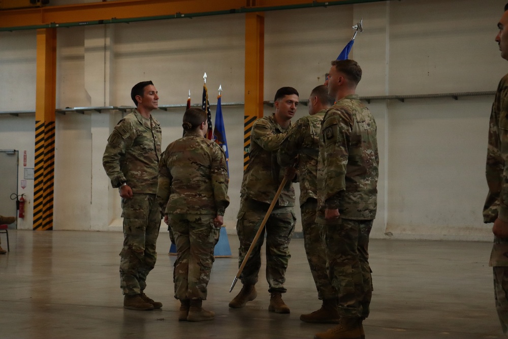 A military ceremony taking place inside what appears to be an aircraft hangar or large military facility. Several personnel in OCP (Operational Camouflage Pattern) uniforms are participating in what appears to be a guidon or flag transfer ceremony, a common practice during change-of-command events. The presence of a unit guidon (blue flag) and the American flag in the background supports this interpretation.