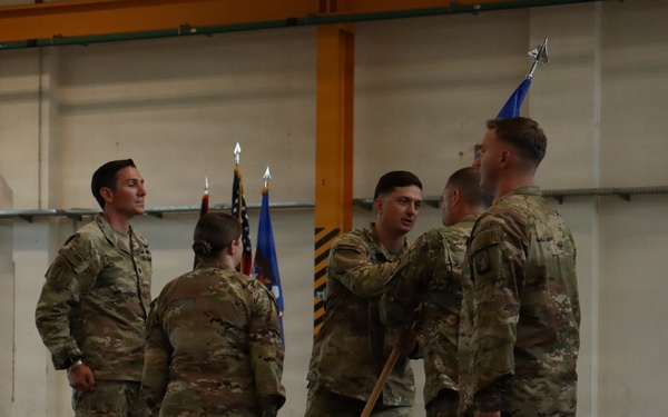 A military ceremony taking place inside what appears to be an aircraft hangar or large military facility. Several personnel in OCP (Operational Camouflage Pattern) uniforms are participating in what appears to be a guidon or flag transfer ceremony, a common practice during change-of-command events. The presence of a unit guidon (blue flag) and the American flag in the background supports this interpretation.