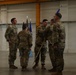 A military ceremony taking place inside what appears to be an aircraft hangar or large military facility. Several personnel in OCP (Operational Camouflage Pattern) uniforms are participating in what appears to be a guidon or flag transfer ceremony, a common practice during change-of-command events. The presence of a unit guidon (blue flag) and the American flag in the background supports this interpretation.