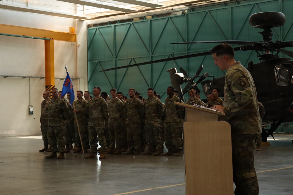A military ceremony is taking place inside an aviation hangar, with a uniformed soldier speaking at a podium before a formation of troops. A military helicopter, appearing to be an AH-64 Apache, is visible in the background. A guidon (unit flag) in blue and orange is being carried by one of the soldiers in the formation.