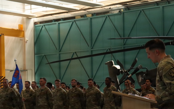 A military ceremony is taking place inside an aviation hangar, with a uniformed soldier speaking at a podium before a formation of troops. A military helicopter, appearing to be an AH-64 Apache, is visible in the background. A guidon (unit flag) in blue and orange is being carried by one of the soldiers in the formation.