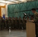A military ceremony is taking place inside an aviation hangar, with a uniformed soldier speaking at a podium before a formation of troops. A military helicopter, appearing to be an AH-64 Apache, is visible in the background. A guidon (unit flag) in blue and orange is being carried by one of the soldiers in the formation.