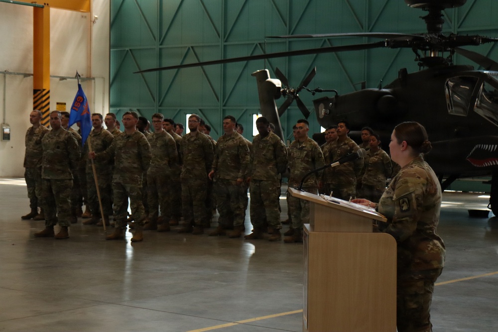 A military ceremony is taking place inside an aircraft hangar, with a female soldier speaking at a podium before a formation of soldiers in camouflage uniforms. A unit guidon (blue flag) is visible being carried by one of the soldiers in formation. An attack helicopter, appearing to be an AH-64 Apache, is visible in the background of the hangar.