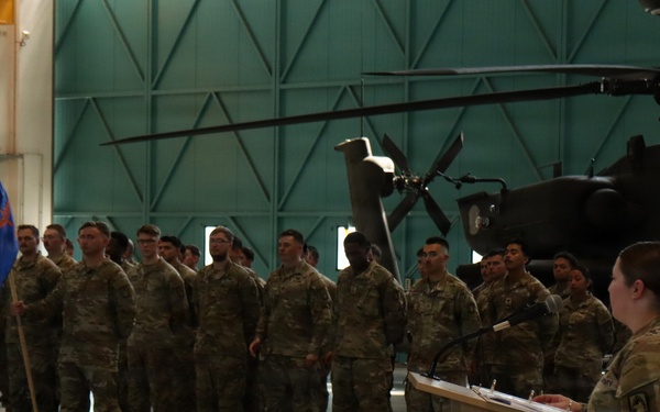 A military ceremony is taking place inside an aircraft hangar, with a female soldier speaking at a podium before a formation of soldiers in camouflage uniforms. A unit guidon (blue flag) is visible being carried by one of the soldiers in formation. An attack helicopter, appearing to be an AH-64 Apache, is visible in the background of the hangar.