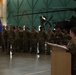 A military ceremony is taking place inside an aircraft hangar, with a female soldier speaking at a podium before a formation of soldiers in camouflage uniforms. A unit guidon (blue flag) is visible being carried by one of the soldiers in formation. An attack helicopter, appearing to be an AH-64 Apache, is visible in the background of the hangar.