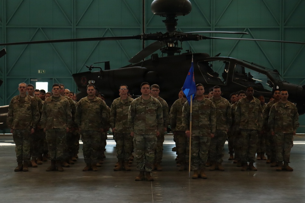 A formation of military personnel in U.S. Army OCP (Operational Camouflage Pattern) uniforms standing at attention inside a large aircraft hangar. A blue unit guidon flag is visible in the center of the formation. An AH-64 Apache attack helicopter is prominently displayed in the background.