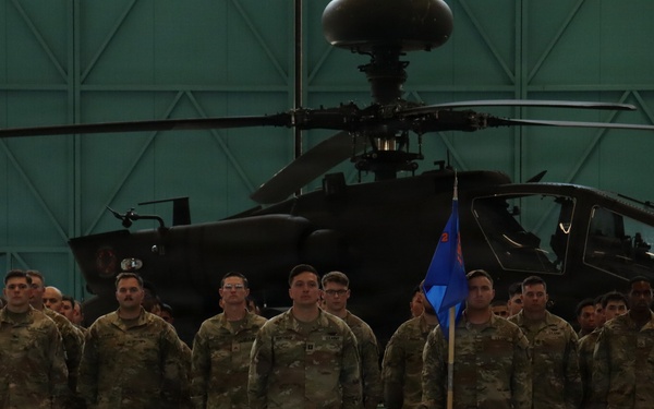 A formation of military personnel in U.S. Army OCP (Operational Camouflage Pattern) uniforms standing at attention inside a large aircraft hangar. A blue unit guidon flag is visible in the center of the formation. An AH-64 Apache attack helicopter is prominently displayed in the background.