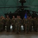 A formation of military personnel in U.S. Army OCP (Operational Camouflage Pattern) uniforms standing at attention inside a large aircraft hangar. A blue unit guidon flag is visible in the center of the formation. An AH-64 Apache attack helicopter is prominently displayed in the background.