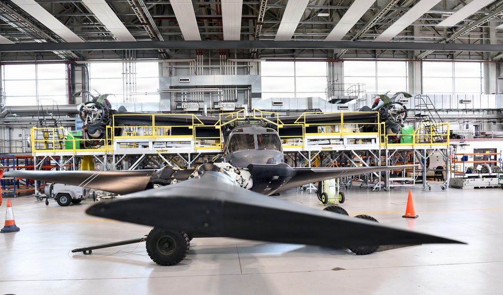 The image shows the interior of a large aircraft manufacturing or maintenance hangar containing multiple aircraft in various stages of assembly or repair. In the foreground is a dark-colored, low-profile aircraft or drone on wheeled landing gear, while behind it sits a larger turboprop aircraft with exposed engines. Yellow industrial scaffolding and work platforms surround the larger aircraft, indicating active maintenance or assembly work.