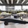 The image shows the interior of a large aircraft manufacturing or maintenance hangar containing multiple aircraft in various stages of assembly or repair. In the foreground is a dark-colored, low-profile aircraft or drone on wheeled landing gear, while behind it sits a larger turboprop aircraft with exposed engines. Yellow industrial scaffolding and work platforms surround the larger aircraft, indicating active maintenance or assembly work.