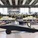 The image shows the interior of a large aircraft manufacturing or maintenance hangar containing multiple aircraft in various stages of assembly or repair. In the foreground is a dark-colored, low-profile aircraft or drone on wheeled landing gear, while behind it sits a larger turboprop aircraft with exposed engines. Yellow industrial scaffolding and work platforms surround the larger aircraft, indicating active maintenance or assembly work.