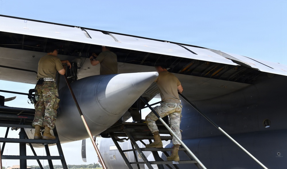 Military personnel in camouflage uniforms are performing maintenance or weapons loading operations on the underside of a large military aircraft. Three individuals are visible working in the bomb bay or weapons bay area, with a large munition or external store visible beneath the wing. The work is being conducted outdoors on what appears to be a flight line or tarmac.