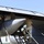 Military personnel in camouflage uniforms are performing maintenance or weapons loading operations on the underside of a large military aircraft. Three individuals are visible working in the bomb bay or weapons bay area, with a large munition or external store visible beneath the wing. The work is being conducted outdoors on what appears to be a flight line or tarmac.
