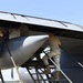 Military personnel in camouflage uniforms are performing maintenance or weapons loading operations on the underside of a large military aircraft. Three individuals are visible working in the bomb bay or weapons bay area, with a large munition or external store visible beneath the wing. The work is being conducted outdoors on what appears to be a flight line or tarmac.
