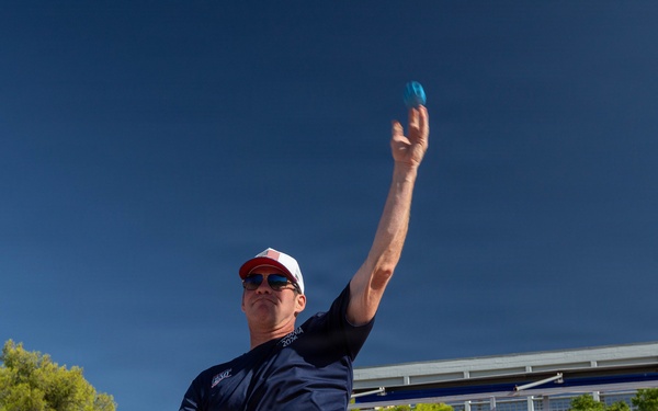 A man in a navy blue t-shirt, white shorts, and a red-brimmed cap is throwing a small blue ball, likely playing pétanque or bocce, at an outdoor sports venue with blue stadium seating. Several onlookers, including individuals in military camouflage uniforms and matching navy team shirts, are watching in the background. The setting appears to be an organized sporting event, possibly a military or international athletic competition.