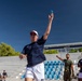 A man in a navy blue t-shirt, white shorts, and a red-brimmed cap is throwing a small blue ball, likely playing pétanque or bocce, at an outdoor sports venue with blue stadium seating. Several onlookers, including individuals in military camouflage uniforms and matching navy team shirts, are watching in the background. The setting appears to be an organized sporting event, possibly a military or international athletic competition.