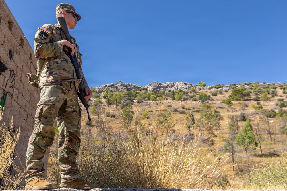 A U.S. Army soldier in full MultiCam (OCP) camouflage uniform stands at a elevated position beside a stone wall, holding a rifle and scanning the surrounding terrain. The soldier wears visible patches including a U.S. flag and what appears to be a NATO patch on the left shoulder. The setting appears to be a semi-arid, hilly landscape with sparse vegetation under a clear blue sky.