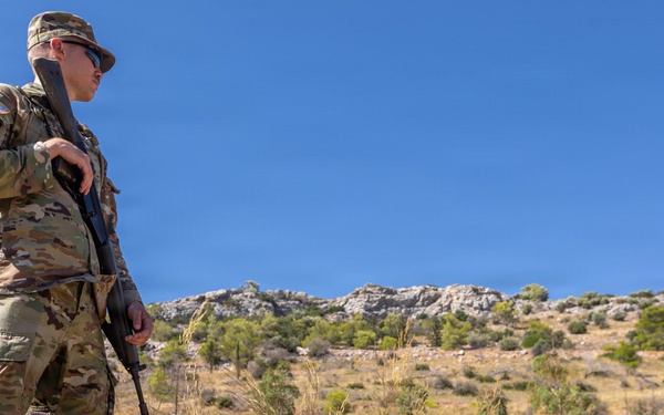 A U.S. Army soldier in full MultiCam (OCP) camouflage uniform stands at a elevated position beside a stone wall, holding a rifle and scanning the surrounding terrain. The soldier wears visible patches including a U.S. flag and what appears to be a NATO patch on the left shoulder. The setting appears to be a semi-arid, hilly landscape with sparse vegetation under a clear blue sky.