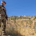 A U.S. Army soldier in full MultiCam (OCP) camouflage uniform stands at a elevated position beside a stone wall, holding a rifle and scanning the surrounding terrain. The soldier wears visible patches including a U.S. flag and what appears to be a NATO patch on the left shoulder. The setting appears to be a semi-arid, hilly landscape with sparse vegetation under a clear blue sky.
