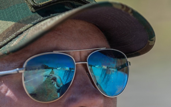 A close-up portrait of a young adult male wearing aviator-style sunglasses with blue mirrored lenses and a camouflage baseball cap. A mustache is prominently visible on his face. The sunglasses reflect a road scene and what appears to be a photographer holding a camera.
