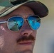 A close-up portrait of a young adult male wearing aviator-style sunglasses with blue mirrored lenses and a camouflage baseball cap. A mustache is prominently visible on his face. The sunglasses reflect a road scene and what appears to be a photographer holding a camera.